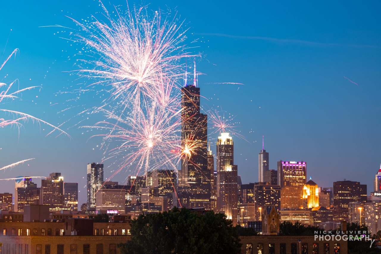 A few fireworks explode in front of the skyline at dusk