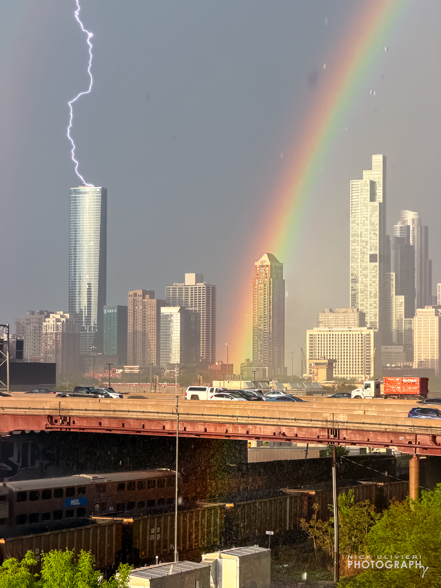 A view of the Chicago skyline after a storm has passed The sun is shin gin creating a vivid rainbow Meanwhile a lignin bolt strikes a skyscraper