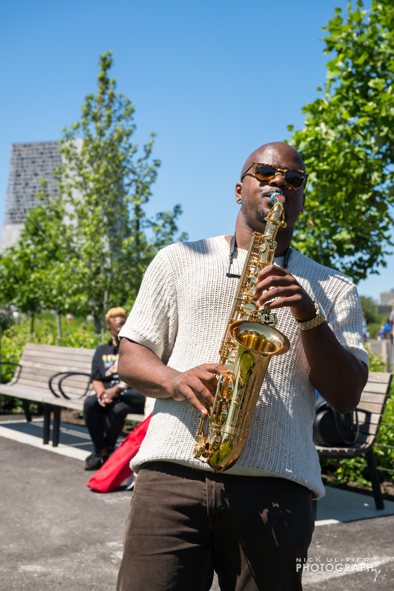 A man plays a saxophone in the park with the Museum tower in the background.