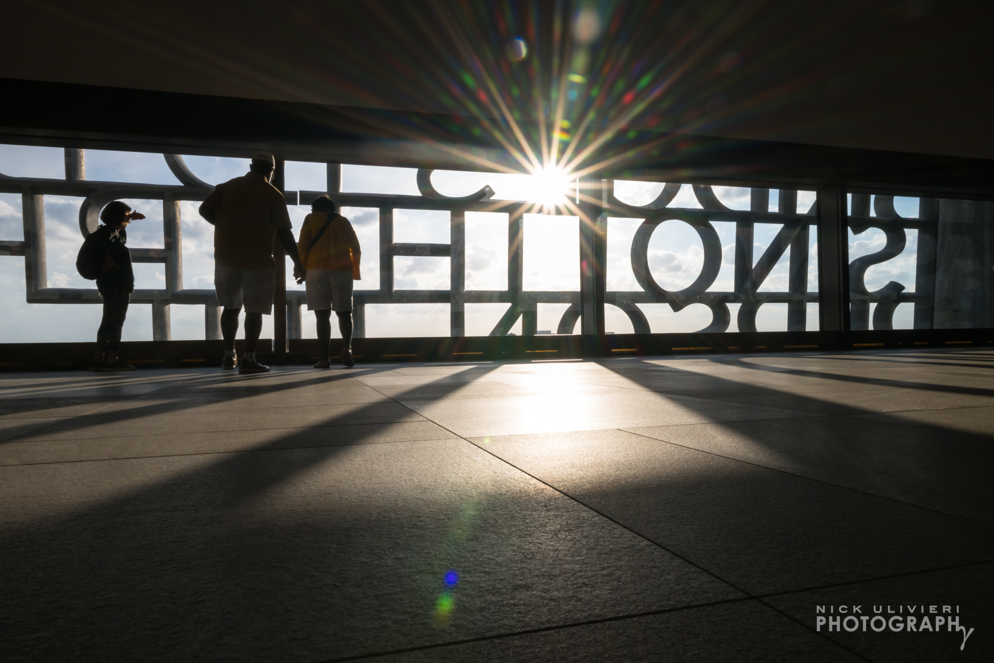 A starburst peeks through the concrete letters of the Sky Room in this low-angle, high-contrast image. Two people are off to the side taking in the view. (public)