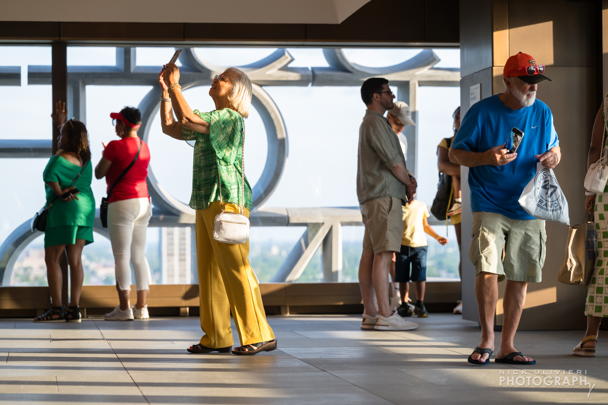 People stand in the Sky Room at the Obama Presidential Center. One woman, wearing a colorful green and yellow out fit looks up to photograph the ceiling (public)