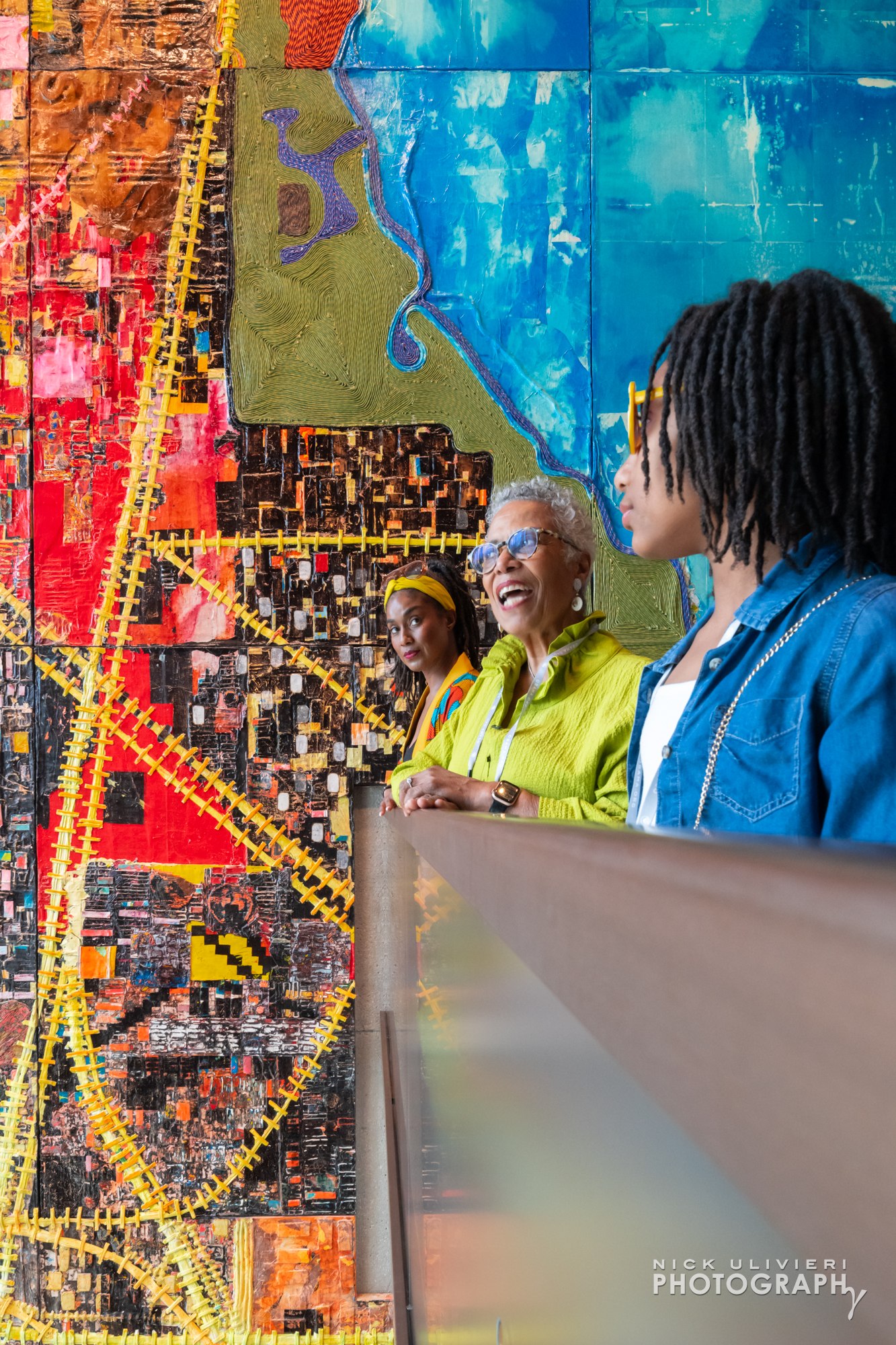 Three generations of women from one family stand near a handrail in the Museum tower looking out and down from the mezzanine. In the background is an extremely colorful, and large piece of art entitled, City of the Big Shoulders by Mark Bradford (public)